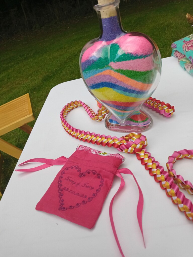 A colourful heart-shaped sandblending vase sits in front of a handfasting ribbon and a ring bag