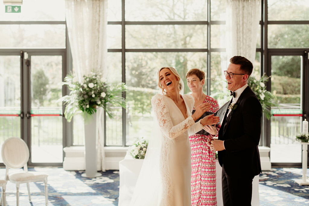 A bride in white and and groom in black stand holding hands. they are laughing. A woman in a patterned dress, the celebrant, stands between them, she is smiling. The bride is looking over her shoulder at unseen guests, her head tilted back in laughter.