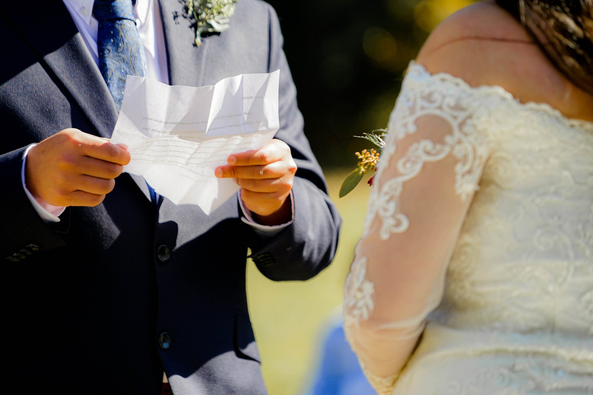 couple renewing their wedding vows man in black suit holding peiece of paper infront of bride.jpg