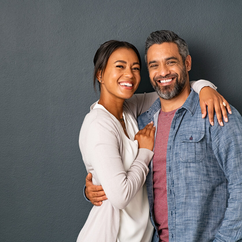 Portrait of happy mid adult couple embracing and looking at camera standing against gray background. Mature indian man in love standing on grey wall while hugging his beautiful hispanic woman. Portarit of carefree mixed race couple with copy space