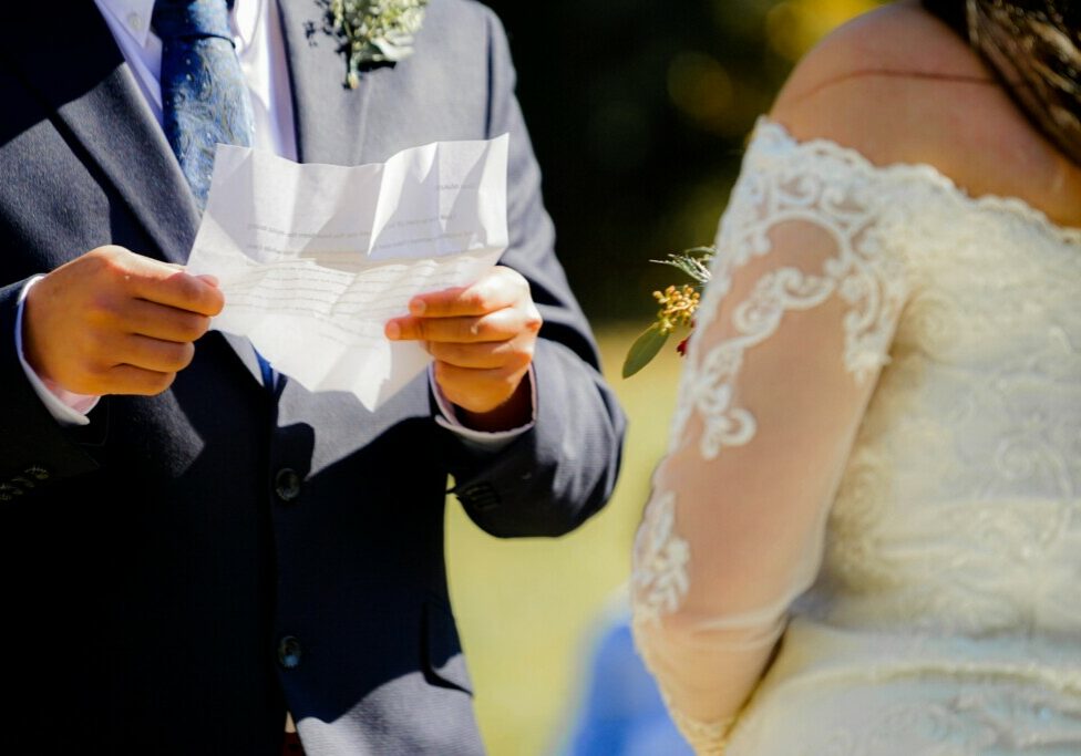 couple renewing their wedding vows man in black suit holding peiece of paper infront of bride.jpg